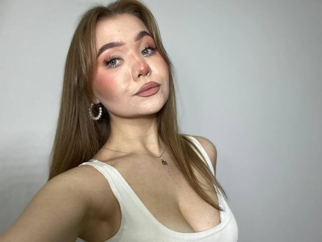 Stacy Yellig ha lunghi capelli castani, occhi azzurri e indossa una canotta bianca che mette in mostra il suo seno. Sta guardando la fotocamera con un'espressione accigliata..