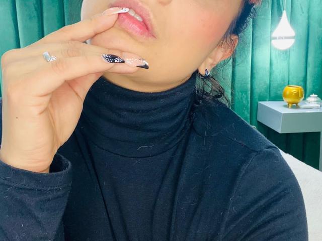 GorgiaFantini's face, black turtleneck, pinkish lips, finger touching mouth, white and black nail with glitter, silver ring, green curtain, and a yellow object in the background.