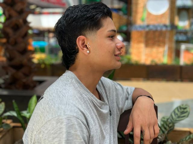JeffTyson, cheveux noirs courts, petits piercings aux oreilles, chemise grise, bras droit posé sur un sac marron, poignet gauche avec un bracelet noir, profil de côté, souriant, en extérieur, en journée..