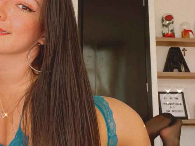 Liliysweet with long brown hair, wearing teal lace bra and black stockings, showing her ass and small tits, large hoop earrings, and a necklace, in a selfie.