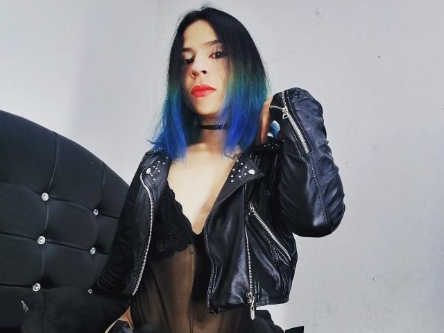 JulesLovets met blauw-groen haar, een zwarte choker, een zwarte leren jas en doorschijnende zwarte lingerie, waarbij haar borsten zichtbaar zijn, draagt rode lippenstift, heeft haar hand in de jas en staat naast een zwarte, gestoffeerde stoel..