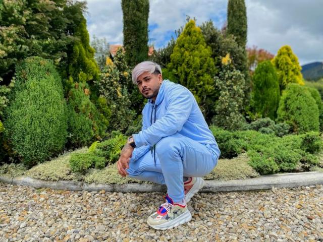VisiI, zittend in een tuin, draagt een lichtblauwe jas en broek, witte sneakers met kleurrijke veters, en grijs haar, met een serieuze blik en een baard..