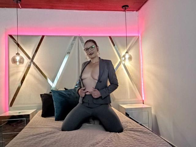 ParisMatur69, in einem grauen Anzug, kniet auf einem Bett, der Anzug ist aufgeknöpft, so dass ihre nackten Brüste zu sehen sind, ihre Hände liegen auf dem Anzug, in einem Raum mit pinkfarbenen LED-Leuchten..