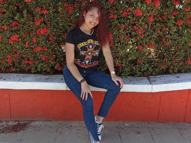 Katrina Bello, com cabelo ruivo encaracolado, sorrindo, vestindo uma camiseta preta com estampa, jeans azuis e tênis azuis, sentada em um banco vermelho e branco, exibindo suas nádegas e seu rosto fofo..