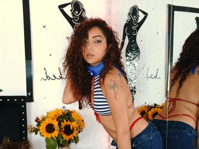Katrina Bello, con cabello rojo rizado, blusa a rayas, corbata azul, parte inferior de traje de baño tipo bikini rojo, tatuaje en el brazo izquierdo, mirando a la cámara, reflejo en un espejo, girasoles, pared blanca con silueta negra..