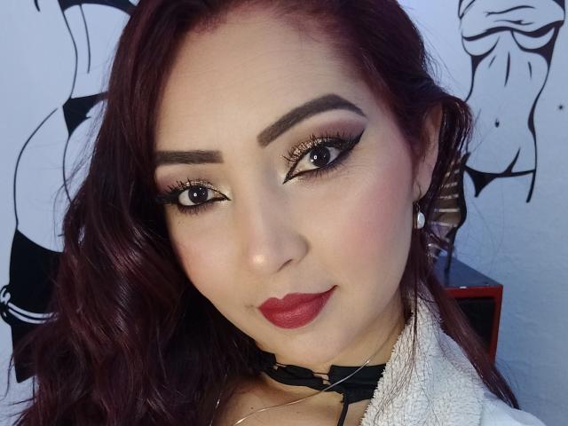 Katrina Bello, avec rouge à lèvres rouge, un collier noir et un eye-liner épais, a des cheveux bruns foncés ondulés, et regarde l'appareil photo avec un léger sourire, montrant ses seins..