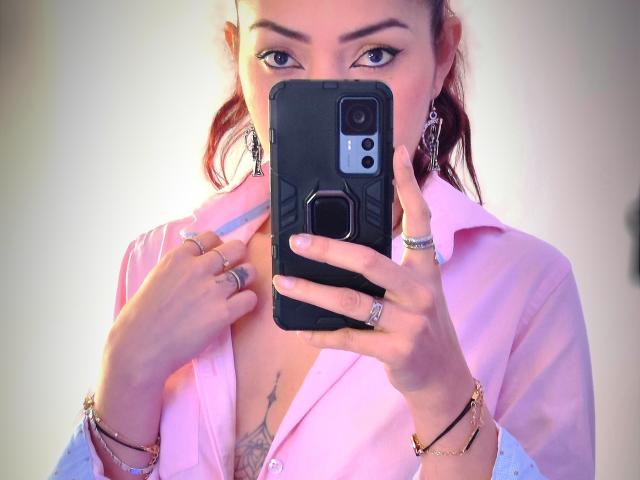 KatrinaBello tirando uma selfie com uma camisa rosa, decote visível, tatuagens, anéis, pulseiras e brincos, segurando um celular preto, unhas rosa e cabelo ruivo em maria-chiquinhas..