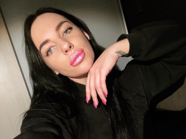StormTammy, con capelli neri, labbra rosa lucide, occhi azzurri, felpa nera, unghie rosa, tatuaggio sul polso destro, appoggiata al braccio, illuminazione soffusa, selfie, sexy, sicura di sé..