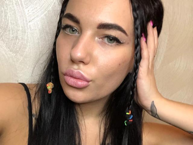 StormTammy heeft zwarte vlechten, roze lippenstift, groene ogen en een tatoeage op haar rechterpols. Haar hand, met roze nagels, raakt haar hoofd aan. Ze draagt een zwarte top..