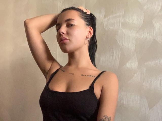 StormTammy, con el pelo negro y húmedo, con una mano en el pelo, vistiendo una camiseta de tirantes negra, con pechos pequeños, tatuajes en el pecho y el brazo, piel morena, expresión de labios fruncidos, de pie contra una pared..