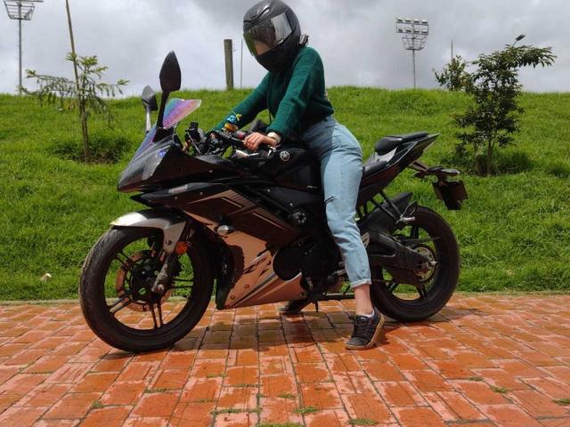 HotAndrea69 com o traseiro à mostra em uma motocicleta preta, vestindo um suéter verde, jeans azuis, um capacete preto e tênis pretos, sentada em uma superfície de tijolos vermelhos..