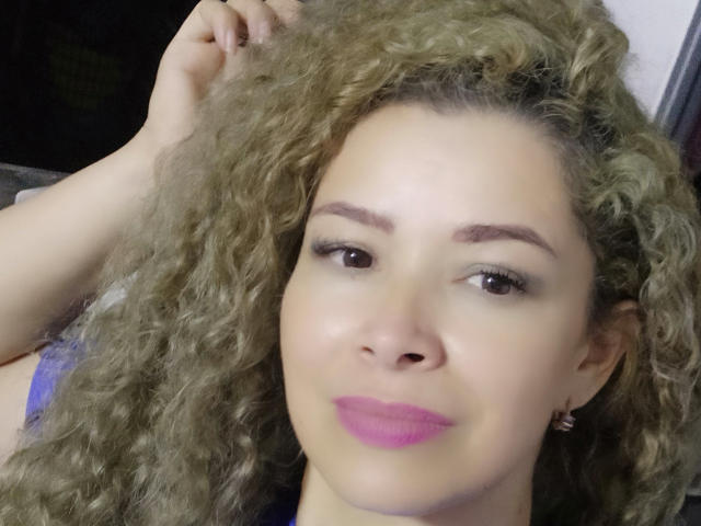 DalilaSuperHot has curly blonde hair, brown eyes, and purple lipstick, with her hand in her hair, wearing a purple top, looking directly at the camera with a slight smile.