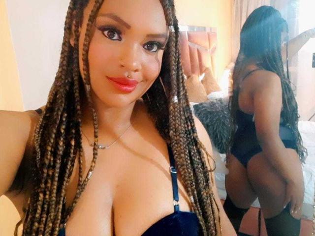 AllyCharlotee with long braided hair, wearing a blue lingerie set, showing her large boobs and big ass in a mirror selfie, holding a phone, smiling with red lipstick.
