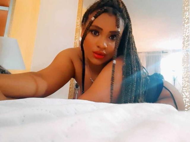 AllyCharlotee with long braids, red lips, and dark skin, lying on a bed, wearing black lingerie, looking at the camera with a sexy, sultry expression.