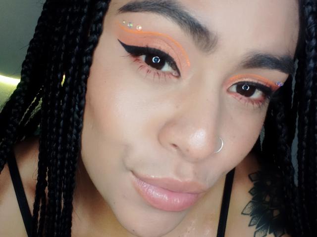 StellaBeckker has braids, a nose ring, and orange eyeliner with black wings, small purple gem on upper eyelid, glossy pink lips, and a tattoo on her right shoulder.