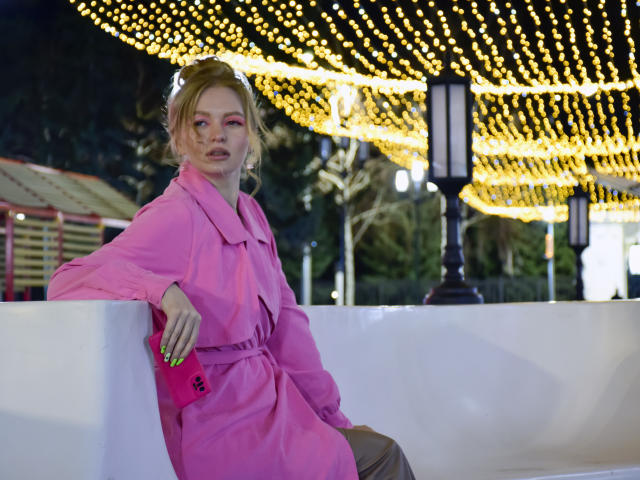 SweetAleksaX with blonde hair, pink coat, neon yellow nails, holding pink phone, leaning on white wall, surrounded by yellow string lights, looking slightly bored.