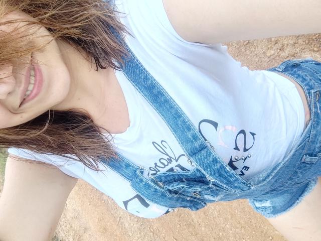 Kkatalina mit welligem, braunem Haar, lächelnd, trägt ein weißes T-Shirt und blaue Jeans-Overalls, wobei ihre Brüste leicht zu sehen sind, draußen auf einer unbefestigten Fläche..