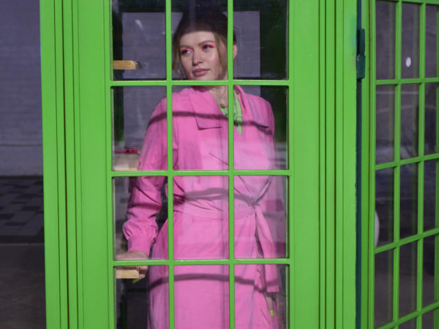 SweetAleksaX stands in a green-framed glass door, wearing a bright pink dress, looking back with pink eye makeup, holding a door handle, showing her pink ass slightly.