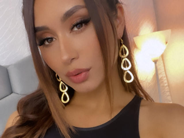 Valeria Klaus, com seus longos cabelos castanhos, vestindo uma blusa preta sem mangas, brincos dourados em formato de gota e batom rosa, tem um visual sensual e sedutor, e está olhando diretamente para a câmera..