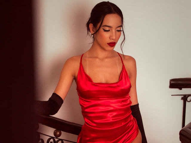 KylieWhite with dark hair, red satin dress, black gloves, and red lipstick, standing by a railing, looking down, showing her tits and slim figure.