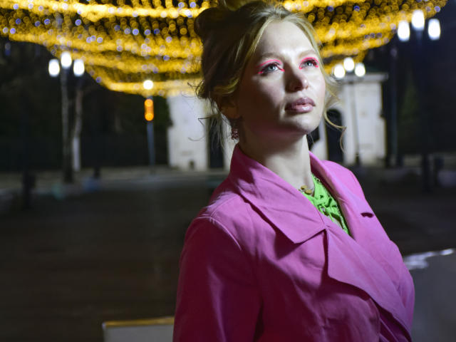 SweetAleksaX con i capelli biondi raccolti in uno chignon, eyeliner rosa, top verde e cappotto rosa, in piedi sotto luci gialle brillanti, che guarda verso l'alto con la bocca leggermente aperta..