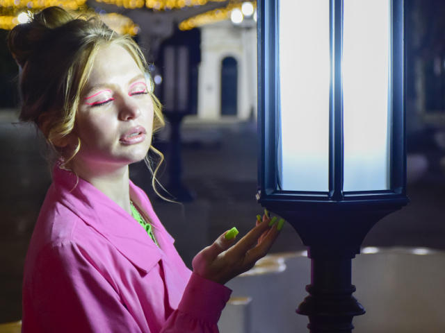 SweetAleksaX, com cabelo loiro preso em um coque, pálpebras rosadas, unhas verde neon e casaco rosa, tocando suavemente um poste de luz brilhante, com os olhos fechados, o rosto iluminado pela luz..