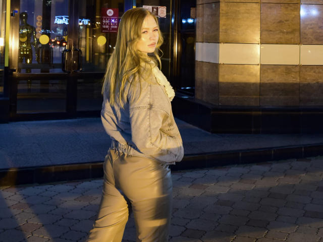 SweetAleksaX with long blonde hair, blue denim jacket, and tan pants, standing on a hexagonal brick sidewalk at night, hands in pockets, looking over shoulder, soft light.