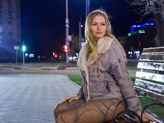 SweetAleksaX with blonde hair, pink eye makeup, and a serious face, wearing a gray fur-lined jacket and brown leather pants, sitting on a metal bench at night.