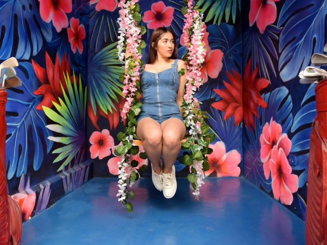 Nico Coleman, com seios médios, sentada em um balanço rosa decorado com flores, vestindo shorts de jeans e tênis brancos, com um fundo colorido de flores. Seu cabelo castanho está solto..