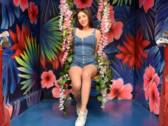 Nico Coleman sentada en un columpio con estampado floral, vistiendo un mono vaquero y zapatillas blancas, con flores rosas y blancas en el columpio, mostrando su trasero y una linda sonrisa..