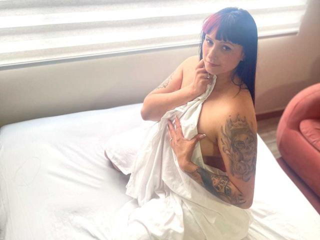 GorgiaFantini with red and black hair, tattoos on her right arm and chest, covering her tits with a white sheet, sitting on a bed, looking at the camera.