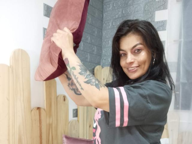 Uma mulher atraente com cabelo preto, sorrindo, segurando um travesseiro vermelho, tem tatuagens no braço direito, vestindo uma camisa preta com listras rosa, em um quarto de madeira..