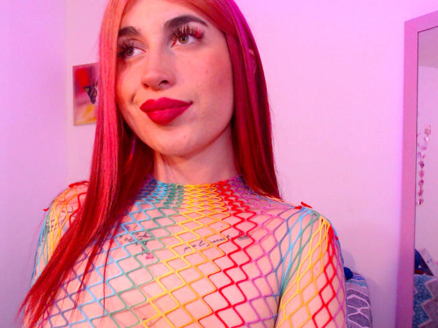 SammyViolet with red hair, wearing a colorful fishnet top, showing her tits through the net, pink lipstick, and a nose ring, smiling at the camera.