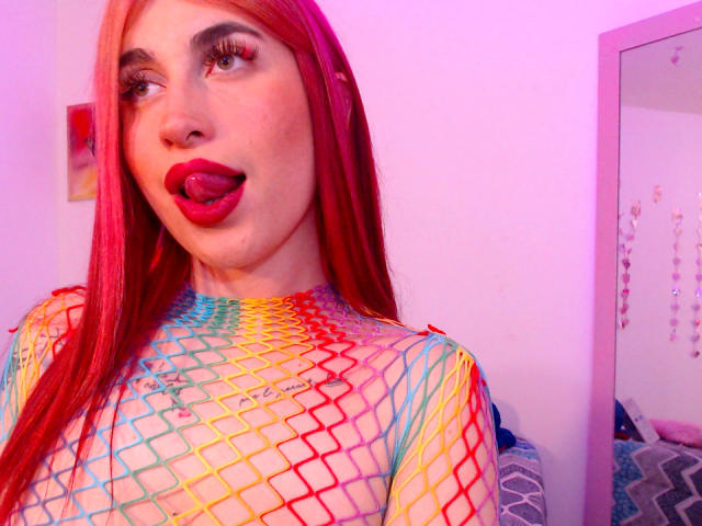 SammyViolet with red hair, tongue out, wearing a rainbow fishnet top, exposing her tits. Her makeup includes bold red lipstick and long lashes. Pink room lighting.