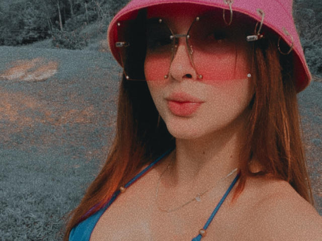 EmilyParkerr with red-tinted glasses, pink bucket hat, nose ring, and blue bikini top, showing her pouty lips and small tits, wearing a necklace in a gritty photo.