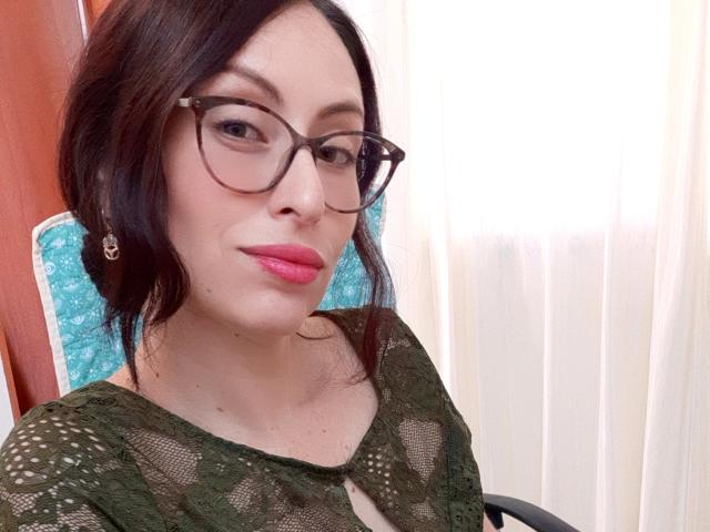 AliceRossi with dark hair, glasses, pink lipstick, and green lace top, showing her cute face and small tits. She's sitting in a chair with a turquoise pattern.