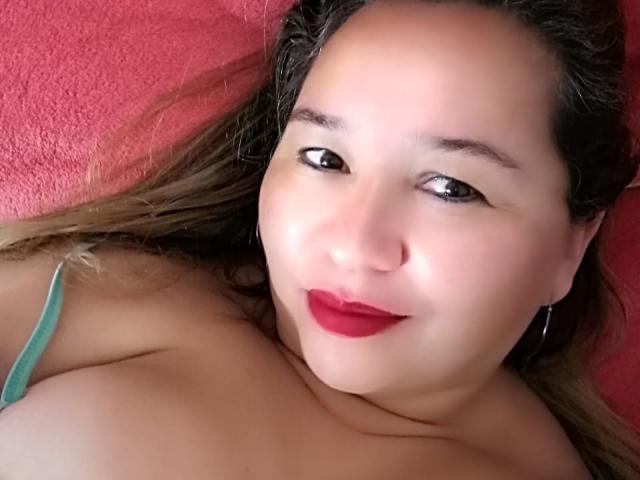 AmbarSimmons with red lipstick, smiling, lying down, showing her tits, wearing a green strap, brown hair, fair skin, and small hoop earrings, against a red wall.