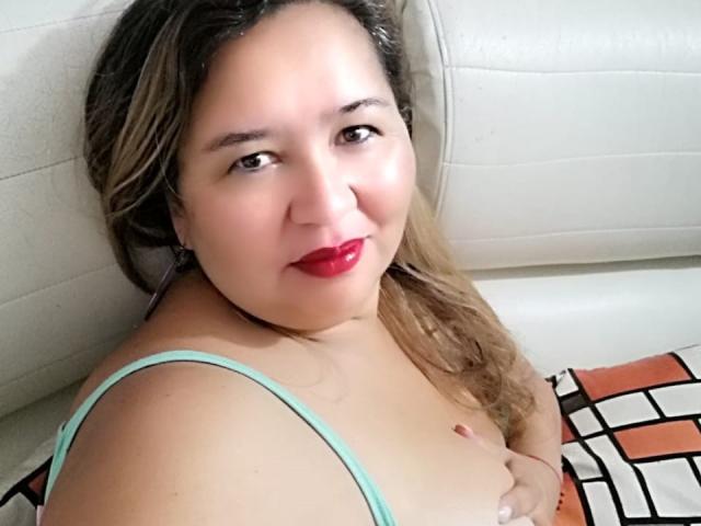 AmbarSimmons with light brown skin, red lipstick, and brown wavy hair, wearing a green strap, covering her tits with her right hand, smiling at the camera.