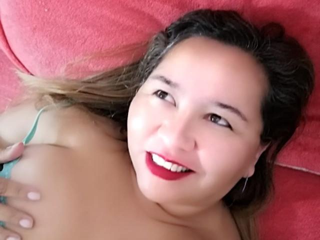 AmbarSimmons, smiling with red lipstick, lying on a pink surface, touching her tits with a green strap, brown eyes, light skin, blonde hair, and hoop earrings, looking up.