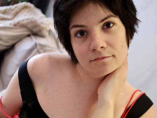 EverlyRays with short black hair, fair skin, and brown eyes, wearing a black top with red straps, resting her chin on her hand, looking at the camera.