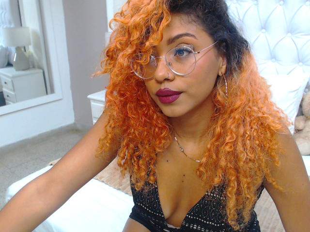 LilithWalker with orange curly hair, wearing glasses, purple lipstick, and a black sparkly top, showing her tits, sitting on a bed, looking at the camera.