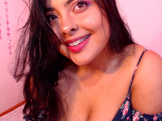 AngelaRoggers with dark hair, pink lipstick, and metal braces, smiling at the camera, wearing a floral off-shoulder top, showing her tits. Pinkish light highlights her face.