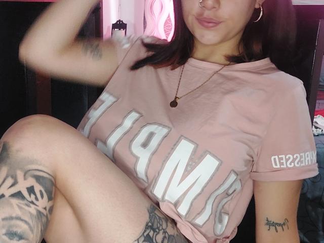 Jazmin und Ryan mit mittelgroßer Brust, tragen ein pinkes T-Shirt mit der Aufschrift "Champs", haben Tattoos an Oberschenkel und Arm, tragen Creolen, eine Nasenring, eine Halskette und haben ein leichtes Lächeln, auf einem Selfie..