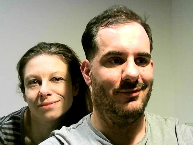 JouwNaughtyKas met borsten en een glimlach, leunt tegen het gezicht van YourNaughtyKas, die een baard en een grijns heeft, in een selfie voor een neutrale grijze muur..