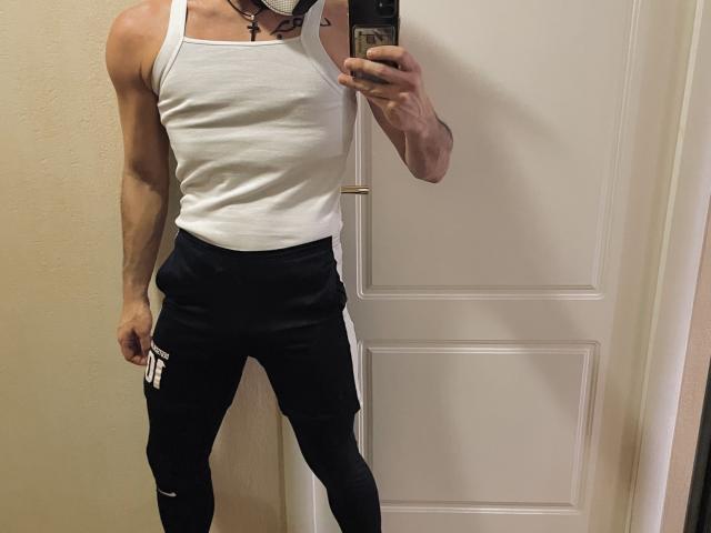 Zamil, com uma estrutura muscular, vestindo uma regata branca e leggings pretas da Nike, segurando um celular em um selfie no espelho, mostrando um colar com uma cruz e uma máscara..