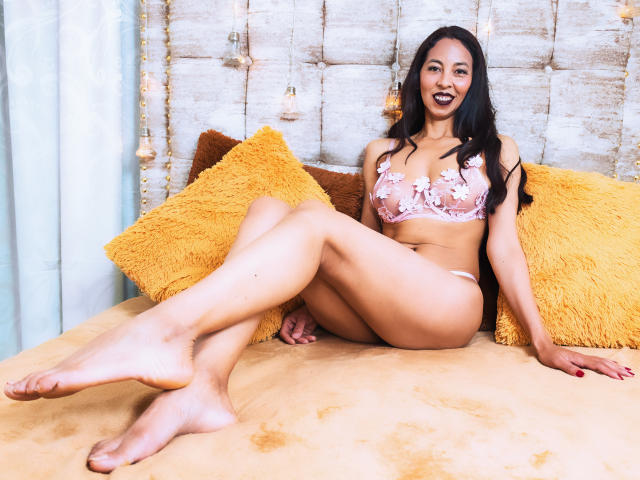 RosalindaCortez, with long black hair, pink floral lingerie, and medium tits, sits on a bed with yellow pillows, legs crossed, smiling with her mouth open.