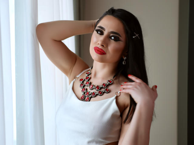 Ema: Ragazza con capelli scuri, rossetto rosso e canotta bianca, con una mano tra i capelli, indossa una collana con una gemma rossa, tiene i capelli, unghie rosse, guarda la fotocamera, sexy, sicura di sé..