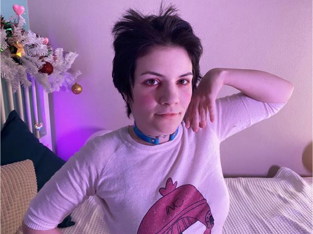 EverlyRays has short black hair, pale skin, and a blue choker. She's wearing a white sweater with a pink hat graphic, resting her arm on a pillow.