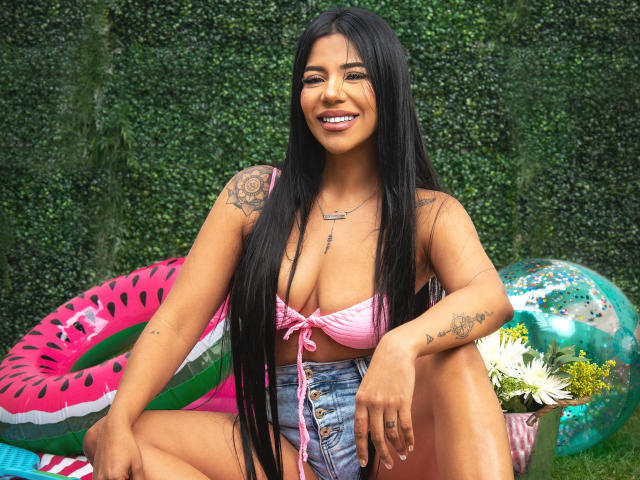 BellaIvanov, com cabelos pretos longos, pele bronzeada e tatuagens, sentada ao ar livre, vestindo um top de biquíni rosa, shorts jeans com botões e um sorriso, com balões de água e flores..