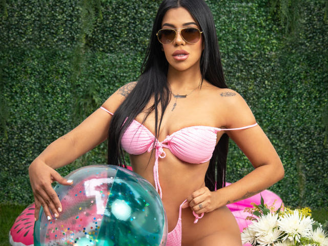 BellaIvanov, avec de longs cheveux noirs, une peau bronzée, portant un bikini rose, tenant un ballon transparent, des lunettes de soleil, des tatouages sur les épaules, assise à l'extérieur, avec des fleurs en bas à droite..